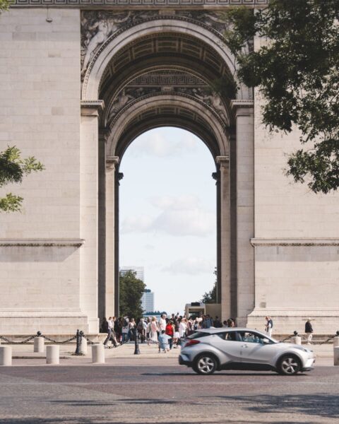 A bustling scene at the Arc de Triomphe with visitors and passing cars captured in daylight.