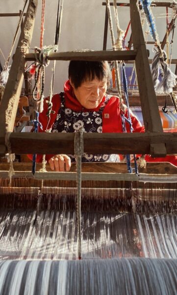 Elderly woman in red weaves on a traditional wooden loom in sunlight.