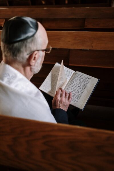 An elderly man with a kippah reads a religious book in a synagogue, wearing a tallit.