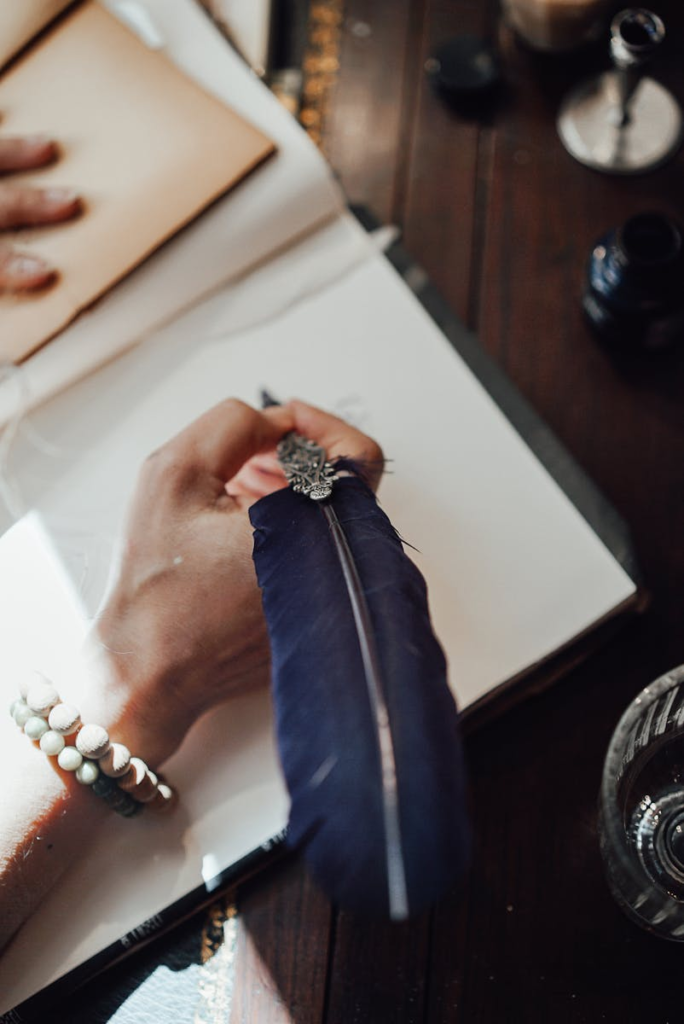 High angle of crop anonymous female author with feather and blank notebook at desk in sun ray